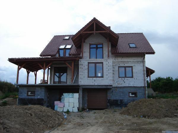 Dom w trakcie budowy z drewnianą konstrukcją dachu i zadaszonym tarasem. Widoczne bloczki betonowe, dachówka i ziemia. Częściowo wykończona elewacja.
