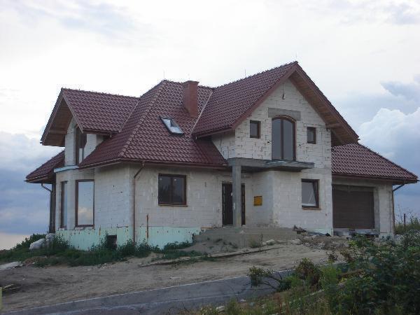 Niedokończony dom jednorodzinny z czerwoną dachówką, widoczne okna z brązowymi ramami, szara elewacja w trakcie budowy, pochmurne niebo w tle.