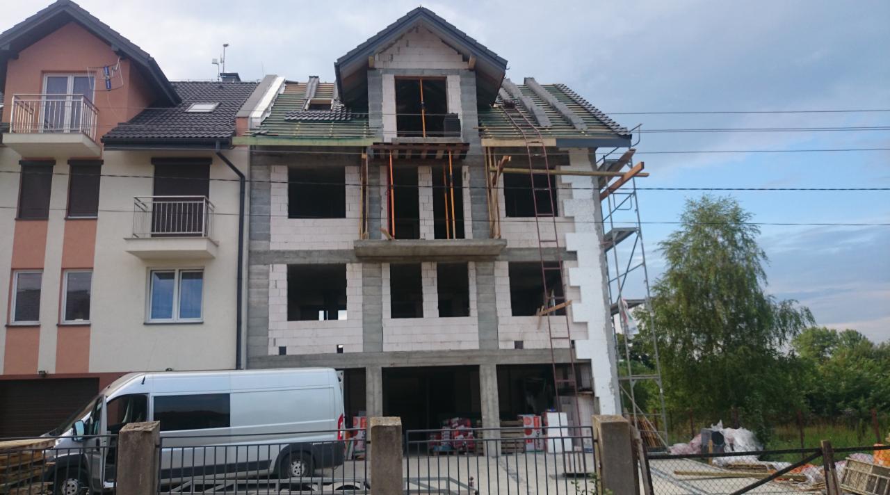 Budynek w trakcie budowy z częściowo położonym dachem i widocznymi rusztowaniami. Na pierwszym planie ogrodzenie i zaparkowany bus.