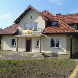 Dwupiętrowy dom jednorodzinny z beżową elewacją, brązowym dachem i numerem 33, z balkonem na pierwszym piętrze, na którym wisi baner reklamowy.