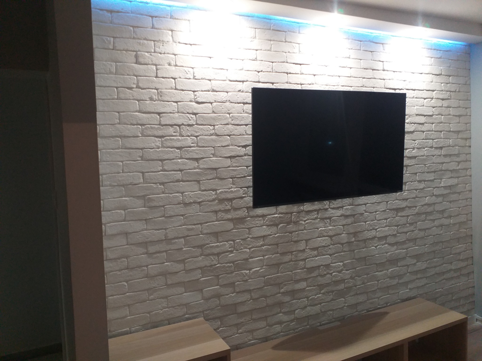Ściana z białej cegły dekoracyjnej z podświetleniem LED u góry, na której wisi czarny telewizor; poniżej widoczny fragment drewnianej szafki RTV.
