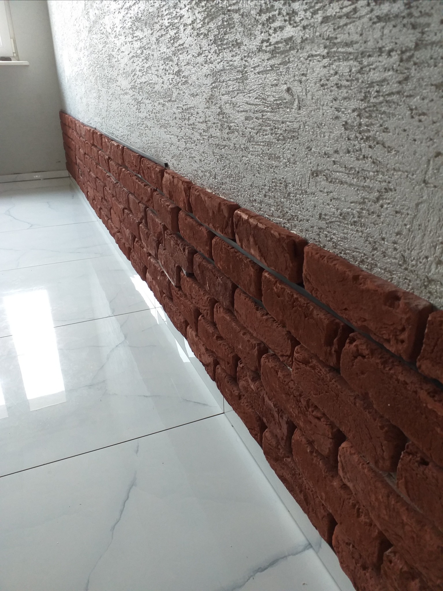 Fragment ściany z dekoracyjną okładziną imitującą czerwoną cegłę, połączoną czarną listwą, na tle ściany z szarą strukturą i marmurowej podłogi z białych płytek.
