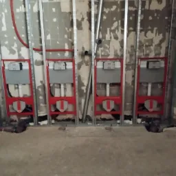 Stelaże podtynkowe WC z czerwonymi ramami zamontowane w metalowej konstrukcji na tle ściany z widocznymi warstwami tynku i instalacjami hydraulicznymi.