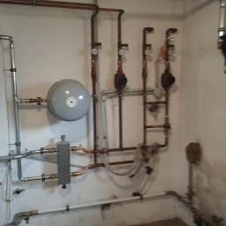 Firma Usługowa Hałys Roman - Instalacja grzewcza z szarym zbiornikiem wyrównawczym, trzema pompami obiegowymi z manometrami, rurami miedzianymi i stalowymi, zamontowana na ścianie w pomieszczeniu technicznym.