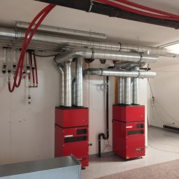 Firma Usługowa Hałys Roman - Dwa czerwone piece grzewcze marki Hoval z podłączonymi srebrnymi rurami wentylacyjnymi i czarnymi rurami instalacji, zamontowane w nowym pomieszczeniu z surowym wykończeniem.