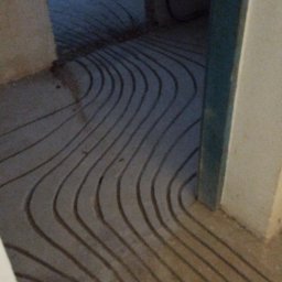 Firma Usługowa Hałys Roman - Ułożone rurki ogrzewania podłogowego w wyżłobionych kanałach w wylewce, widok z poziomu podłogi, na dalszym planie pomieszczenie w trakcie remontu.