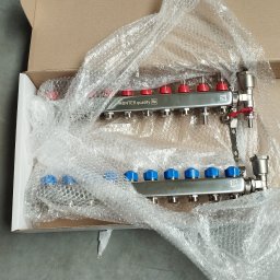 Firma Usługowa Hałys Roman - Rozdzielacz ogrzewania podłogowego Monter Quality w kartonie, zabezpieczony folią bąbelkową. Widoczne czerwone i niebieskie pokrętła regulacyjne. Obok fragment buta.