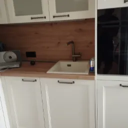 Nowoczesna kuchnia w bieli z drewnianym blatem i baterią przy zlewie. Widoczny fragment płyty indukcyjnej i piekarnika. Minimalistyczny design, ciepłe oświetlenie.