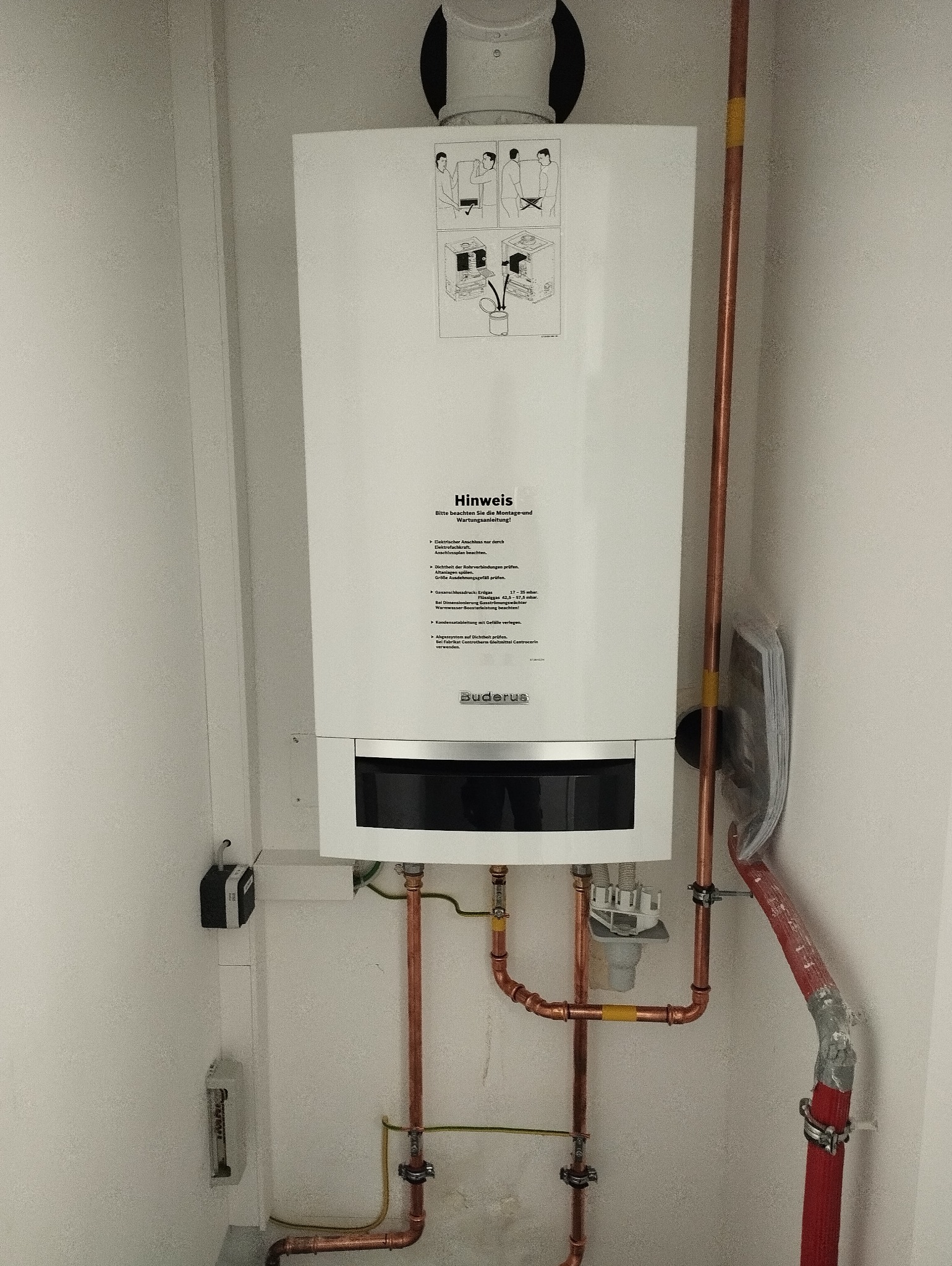 Biały, wiszący piec gazowy marki Buderus z miedzianymi rurami instalacji grzewczej, zamontowany na białej ścianie w pomieszczeniu technicznym.