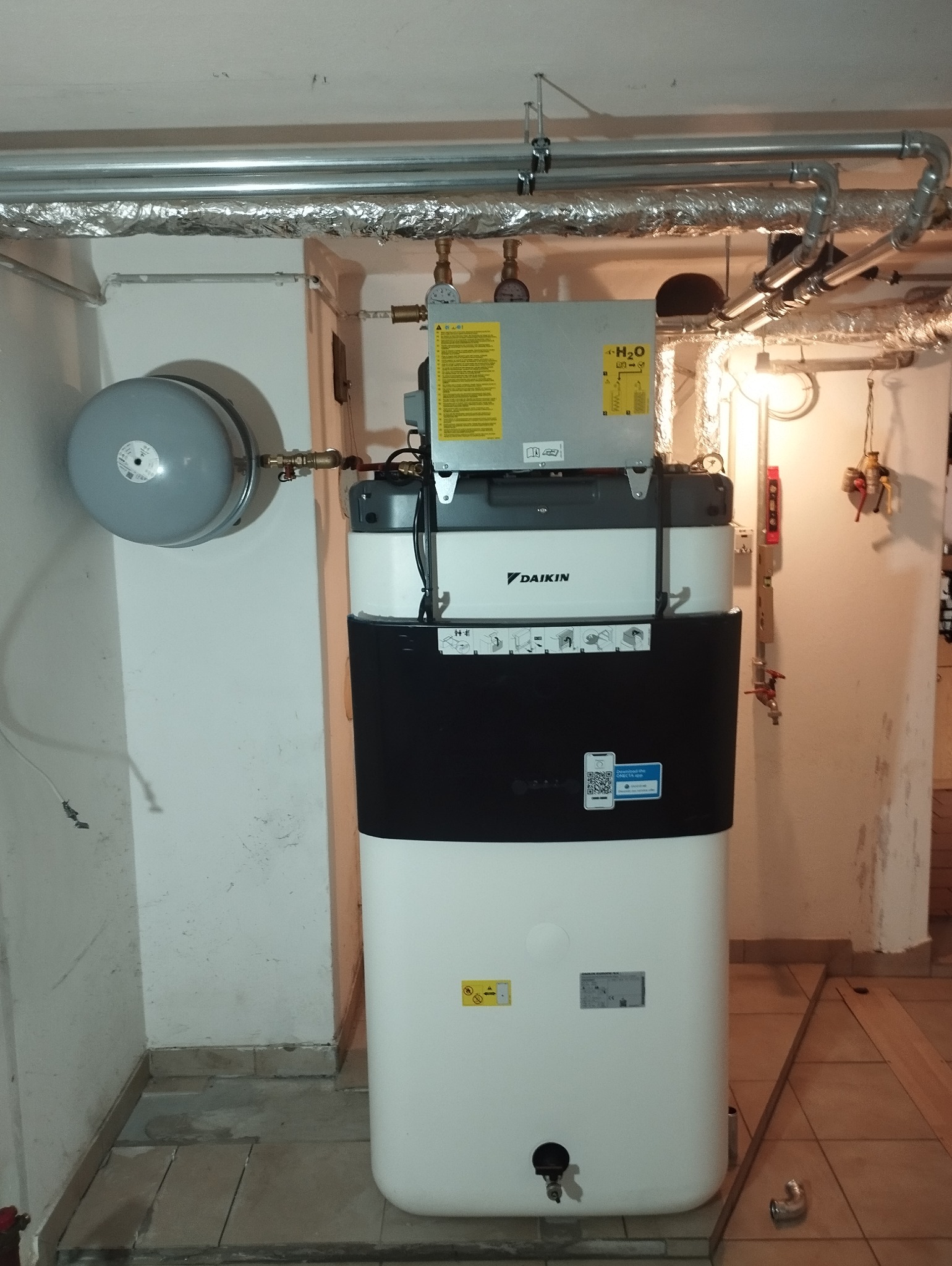 Biało-czarna pompa ciepła Daikin z szarym zbiornikiem wyrównawczym, zainstalowana w piwnicy z widocznymi rurami instalacji grzewczej pod sufitem.
