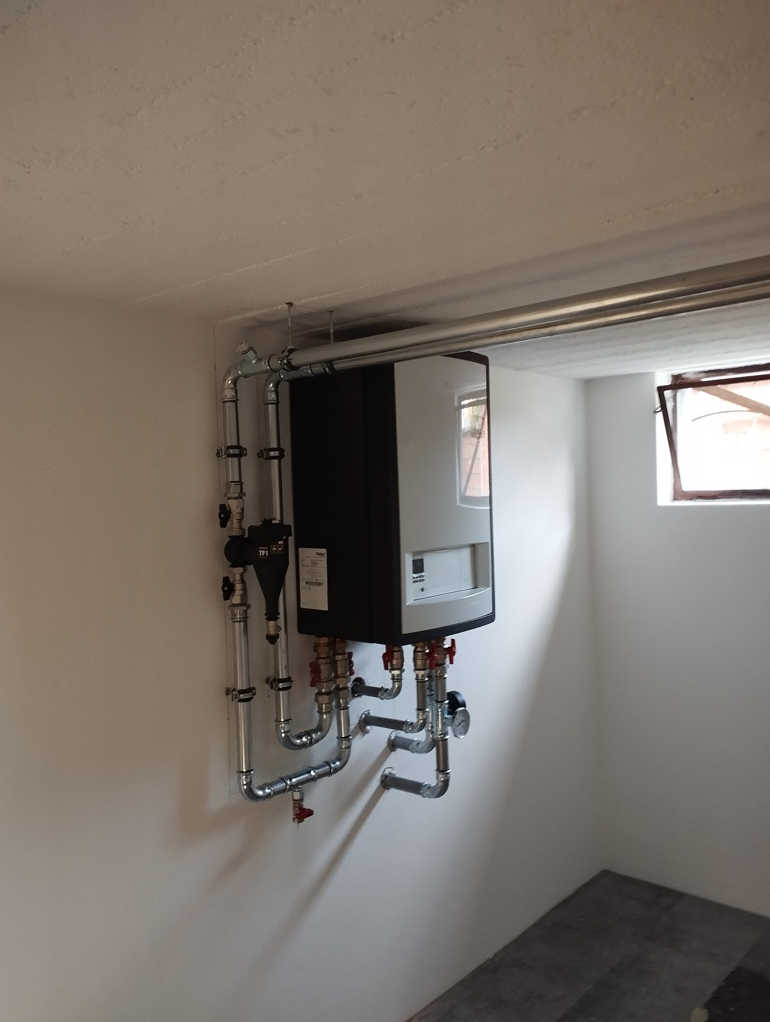 Nowoczesny, wiszący piec grzewczy z czarnymi bokami i białym frontem, podłączony do instalacji z rur chromowanych, zamontowany w pomieszczeniu z małym oknem i betonową podłogą.