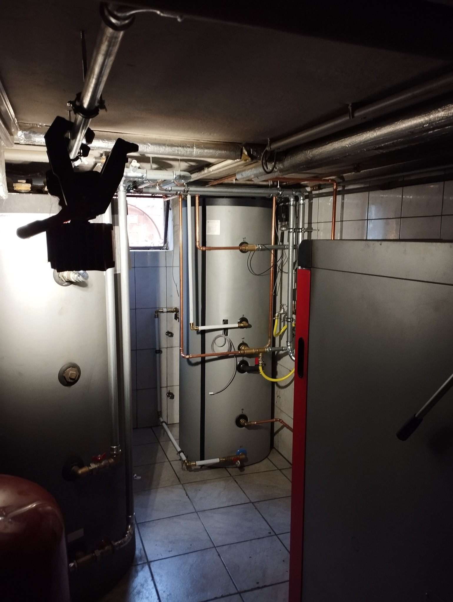 Piwnica z zainstalowanymi zbiornikami na wodę i rurami miedzianymi, widoczne rury pod sufitem i małe okno w tle.