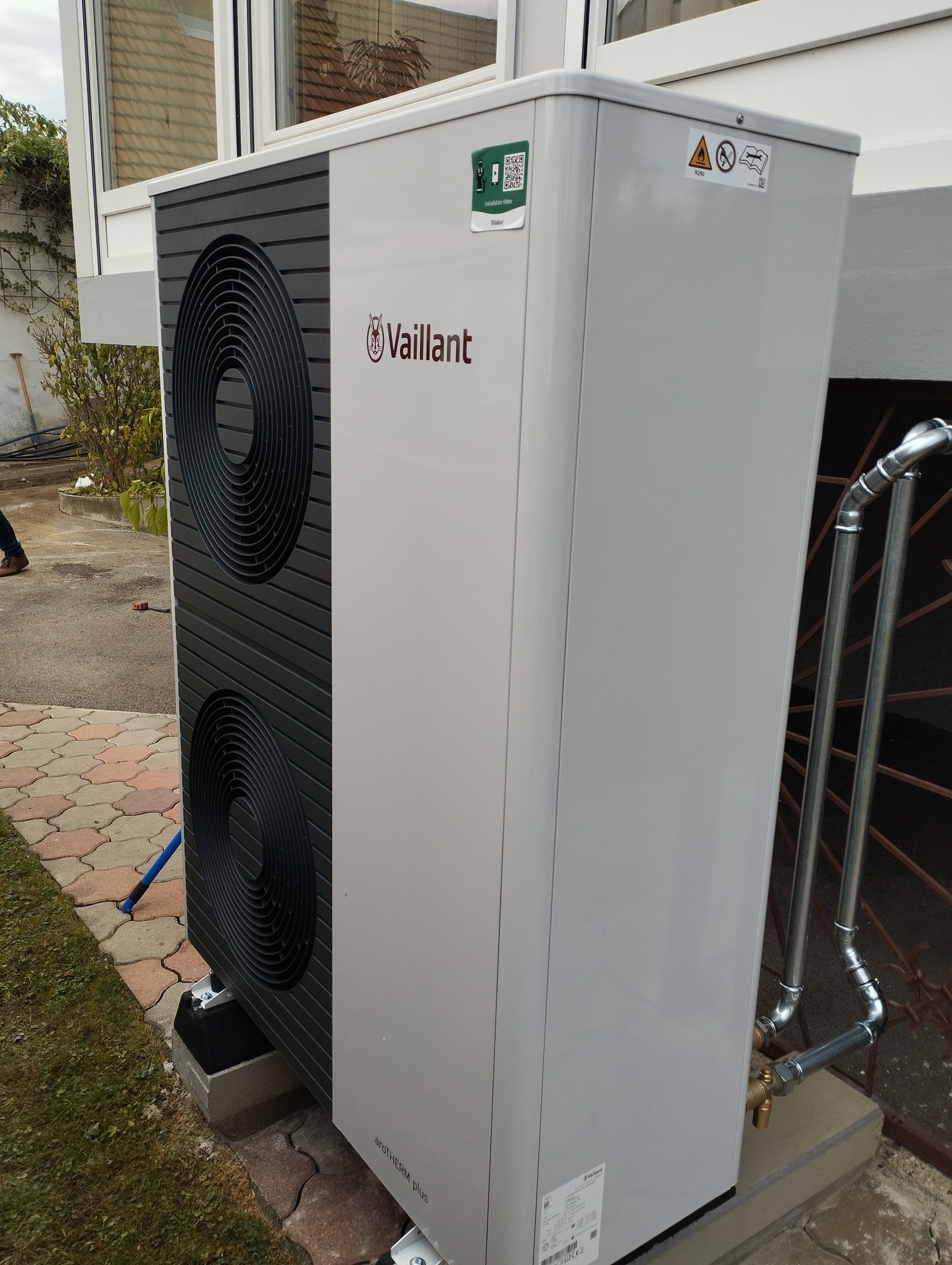 Zewnętrzna jednostka pompy ciepła Vaillant aroTHERM plus, widoczne przyłącza rur, zamontowana na betonowych bloczkach, z naklejkami informacyjnymi i kodem QR.