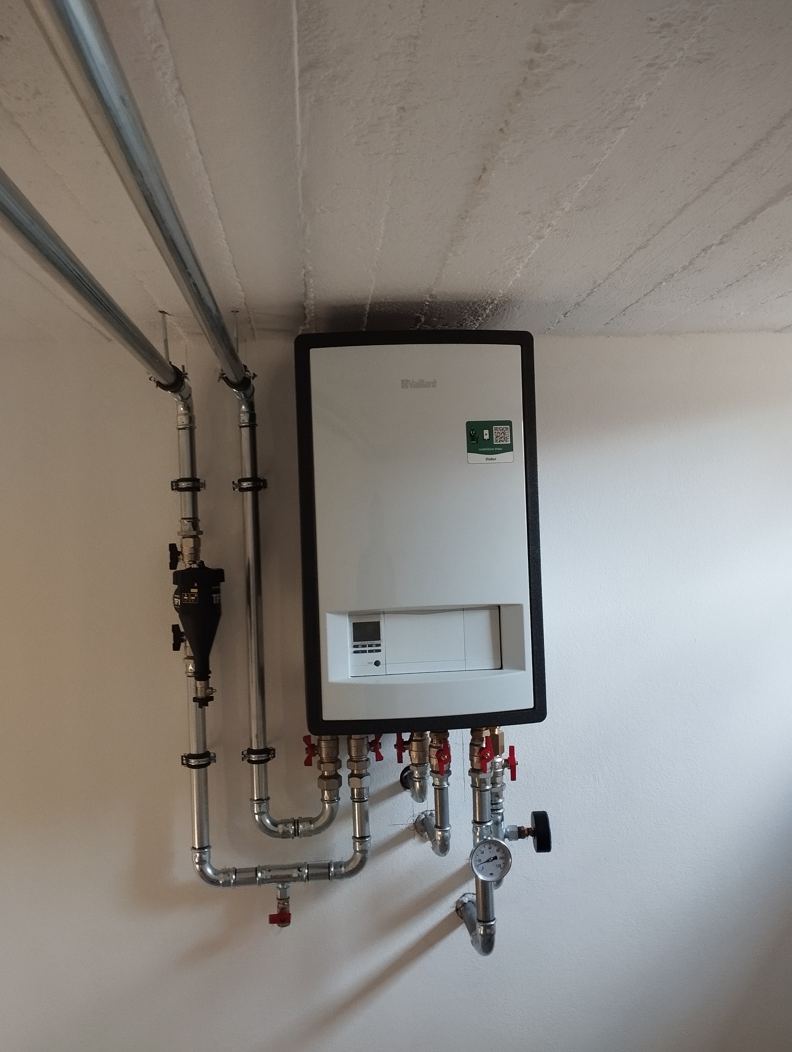 Nowoczesny, biały piec gazowy marki Vaillant z czarną obudową, zamontowany na białej ścianie, z widocznymi rurami, zaworami i manometrem. Na górze widoczne dwie równoległe rury.