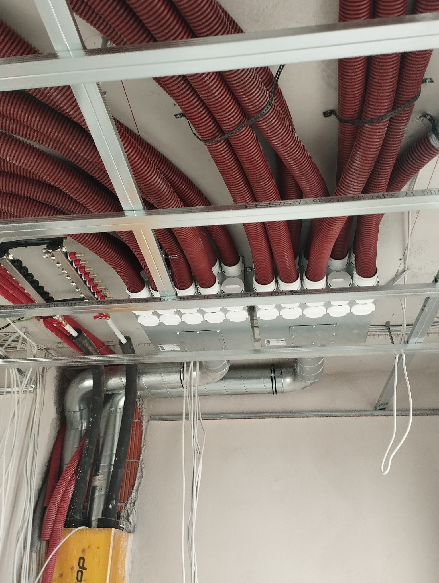 Czerwone karbowane rury instalacyjne poprowadzone pod sufitem podwieszanym, z widocznymi puszkami rozdzielczymi i fragmentami instalacji wentylacyjnej, instalacja w trakcie realizacji.