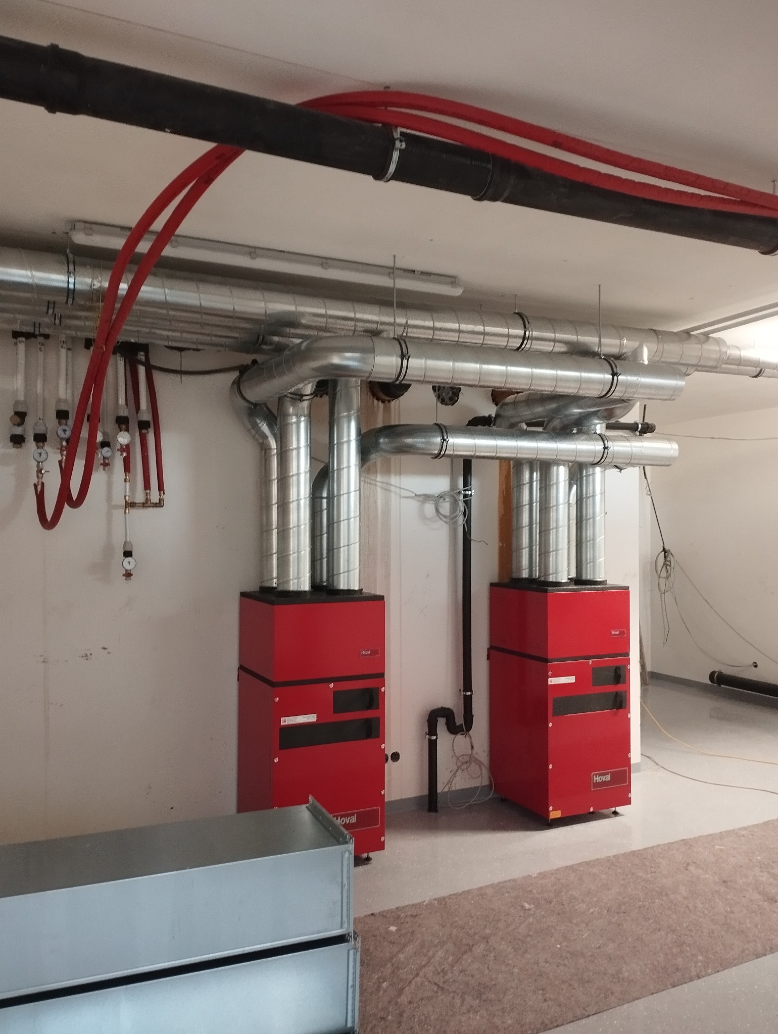 Dwa czerwone piece grzewcze marki Hoval z podłączonymi srebrnymi rurami wentylacyjnymi i czarnymi rurami instalacji, zamontowane w nowym pomieszczeniu z surowym wykończeniem.