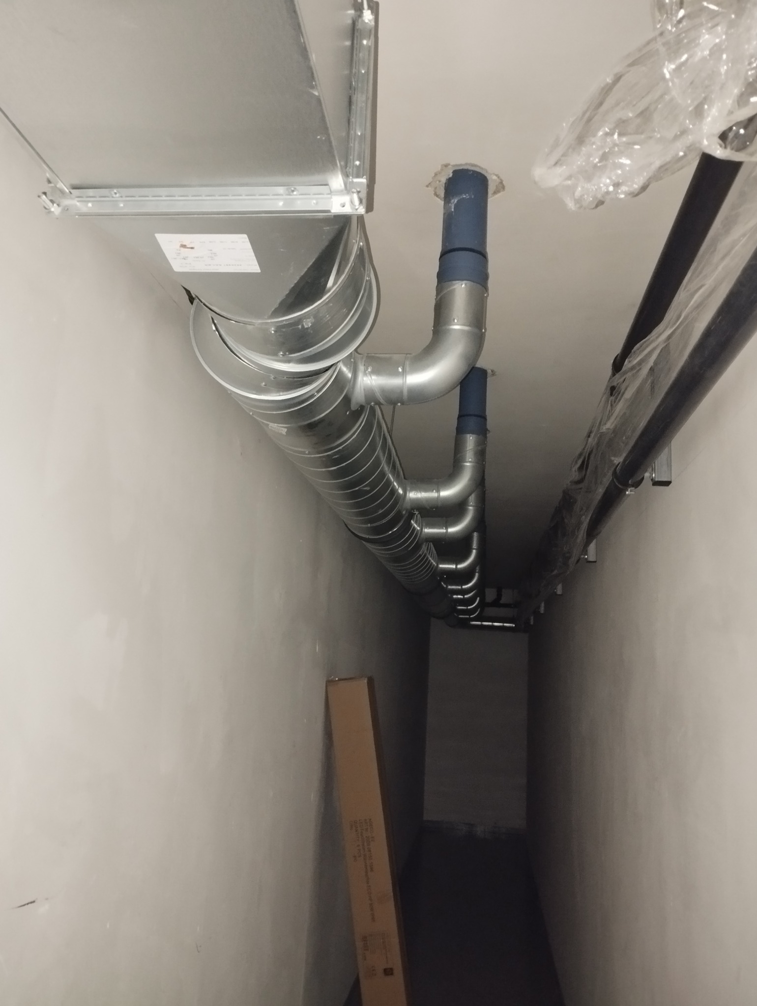 Szereg srebrnych, spiralnych rur wentylacyjnych zamontowanych pod sufitem w korytarzu, z widocznymi elementami mocującymi i niebieskimi łącznikami, obok czarnych rur w folii ochronnej,...