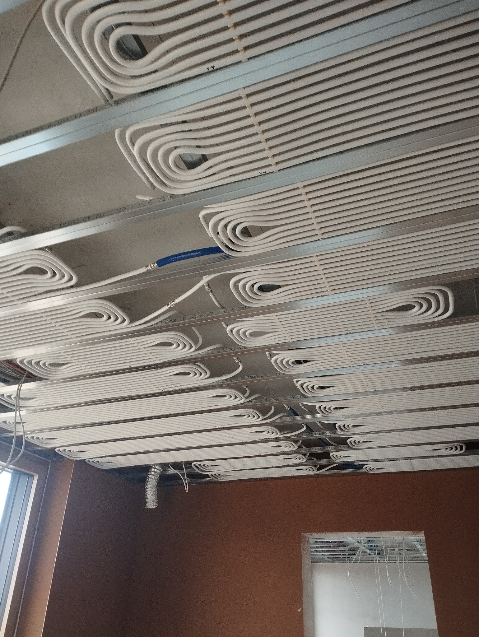 Białe rury ogrzewania podłogowego zamontowane pod sufitem na metalowej konstrukcji, widoczne niebieskie złączki i fragment okna w tle.