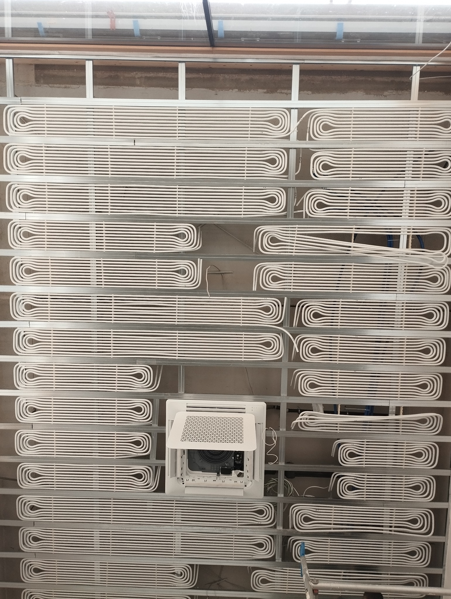 Białe rury ogrzewania podłogowego w metalowej ramie montażowej, widok z dołu, z otwartym panelem klimatyzacji sufitowej w centrum kadru.