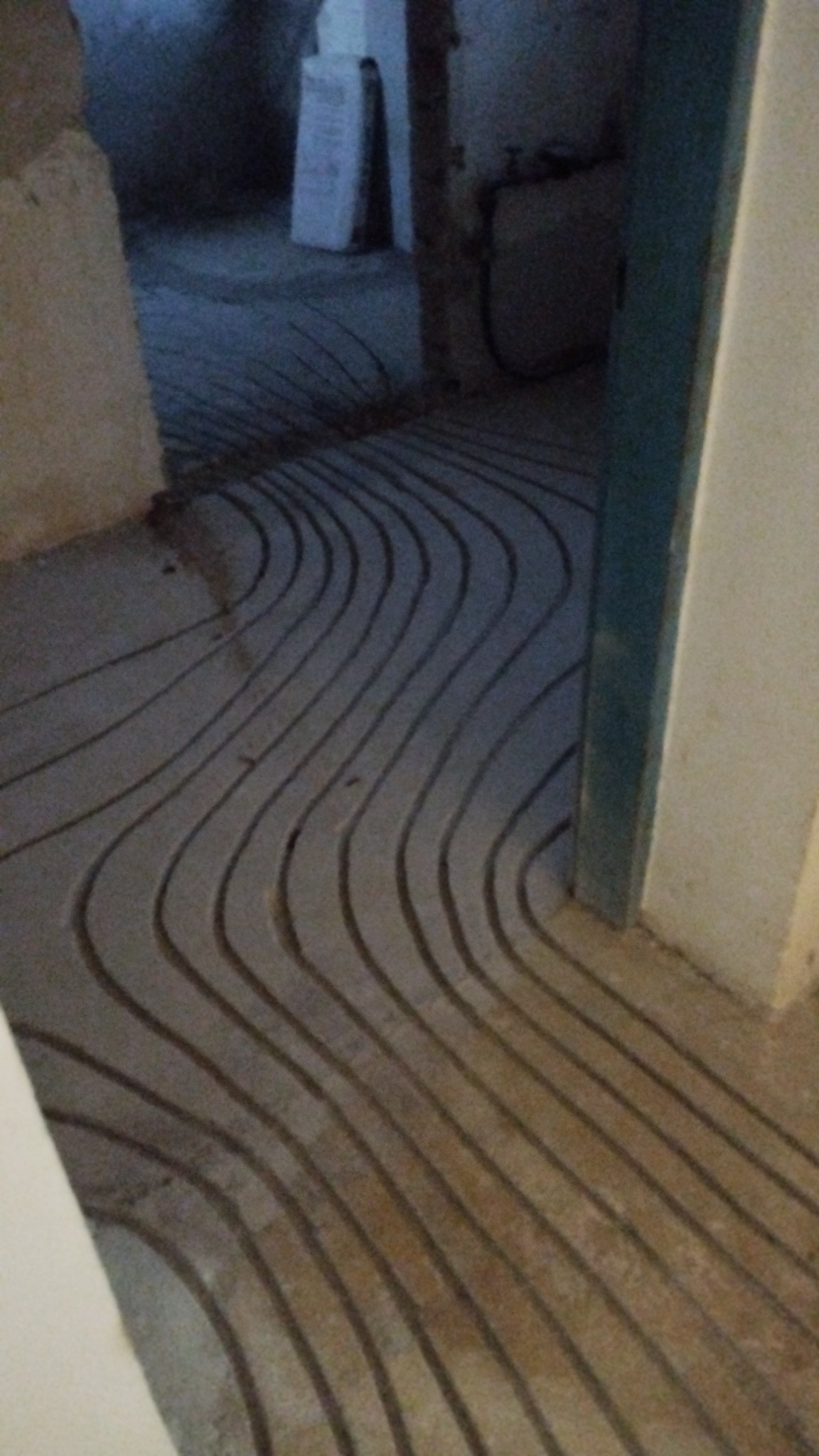 Ułożone rurki ogrzewania podłogowego w wyżłobionych kanałach w wylewce, widok z poziomu podłogi, na dalszym planie pomieszczenie w trakcie remontu.