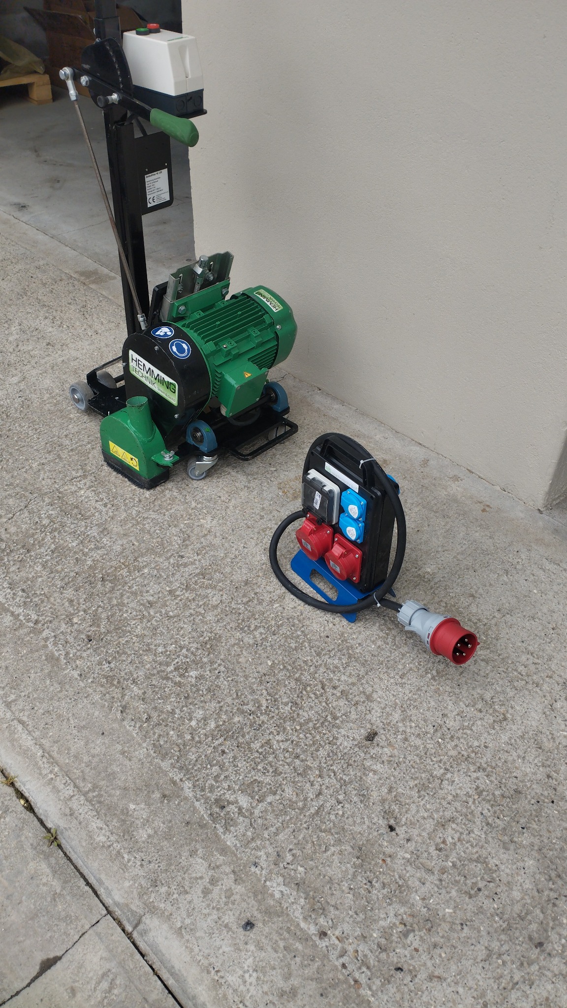 Szlifierka do betonu Hemming i rozdzielnica elektryczna na betonowej posadzce. Widoczne gniazda siłowe i wtyczka. Sprzęt gotowy do użycia.
