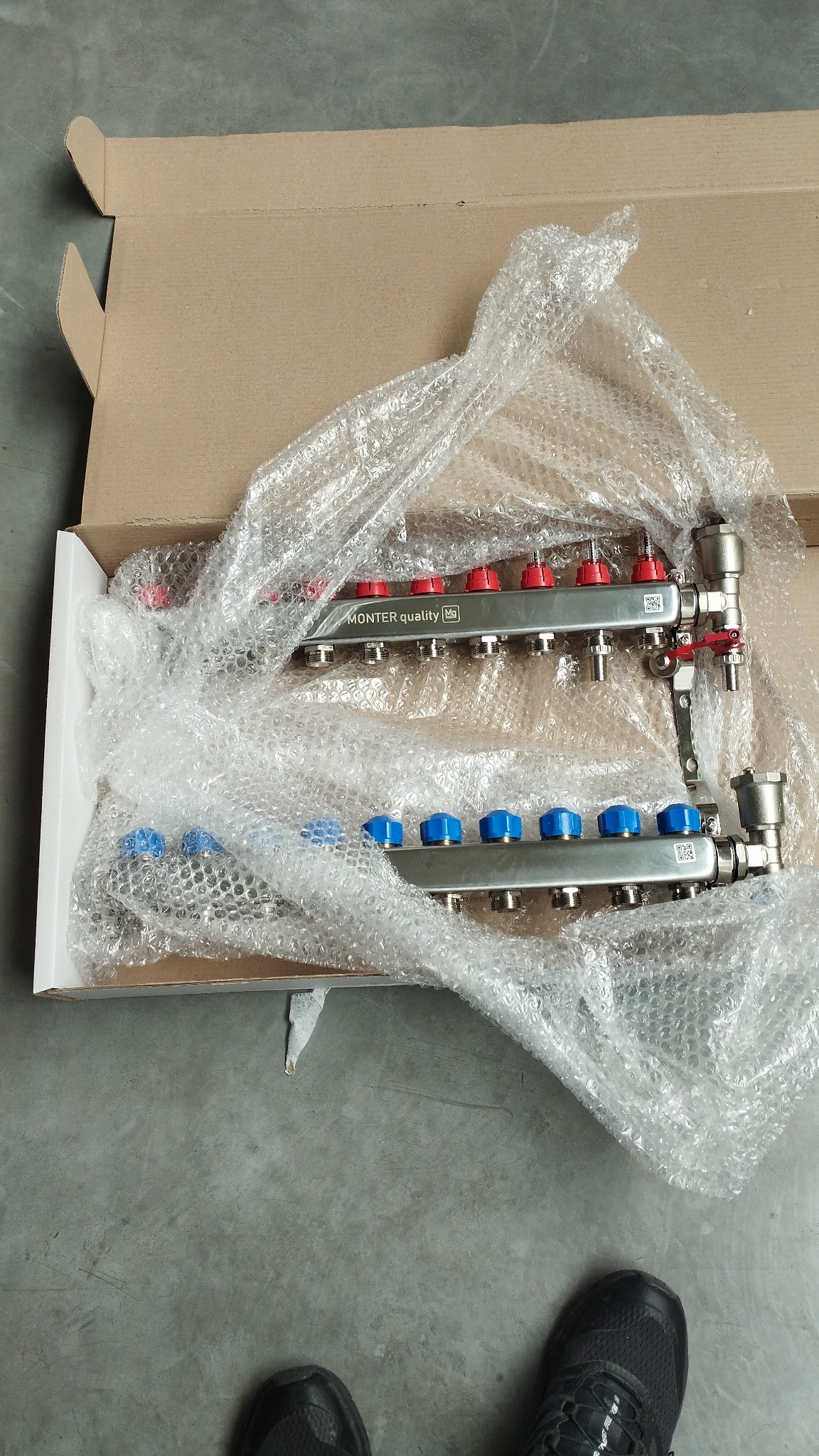 Rozdzielacz ogrzewania podłogowego Monter Quality w kartonie, zabezpieczony folią bąbelkową. Widoczne czerwone i niebieskie pokrętła regulacyjne. Obok fragment buta.