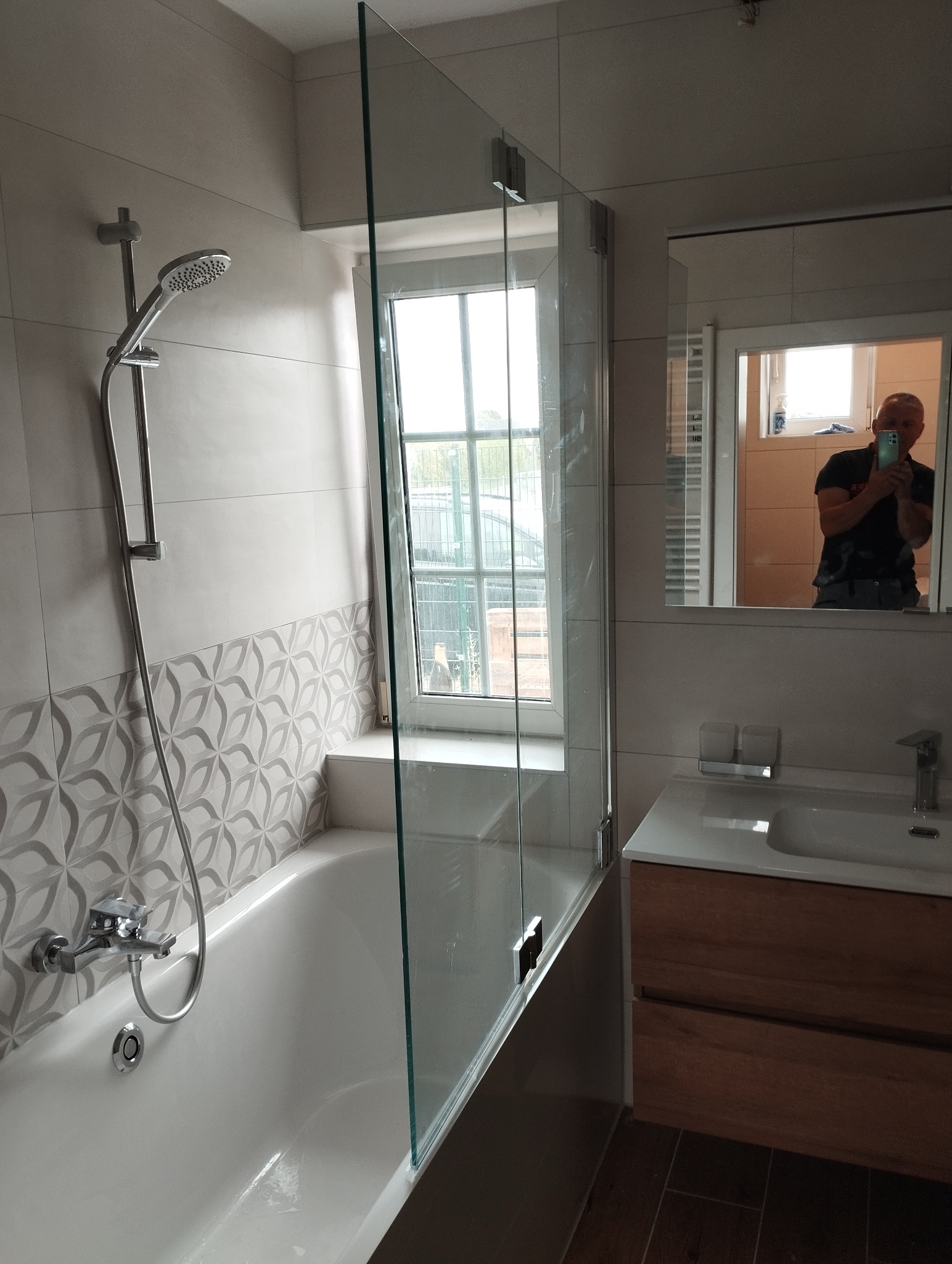 Wykończona łazienka z wanną, szklanym parawanem nawannowym i umywalką z szafką. Widoczne płytki z dekoracyjnym wzorem. W lustrze odbija się osoba robiąca zdjęcie.