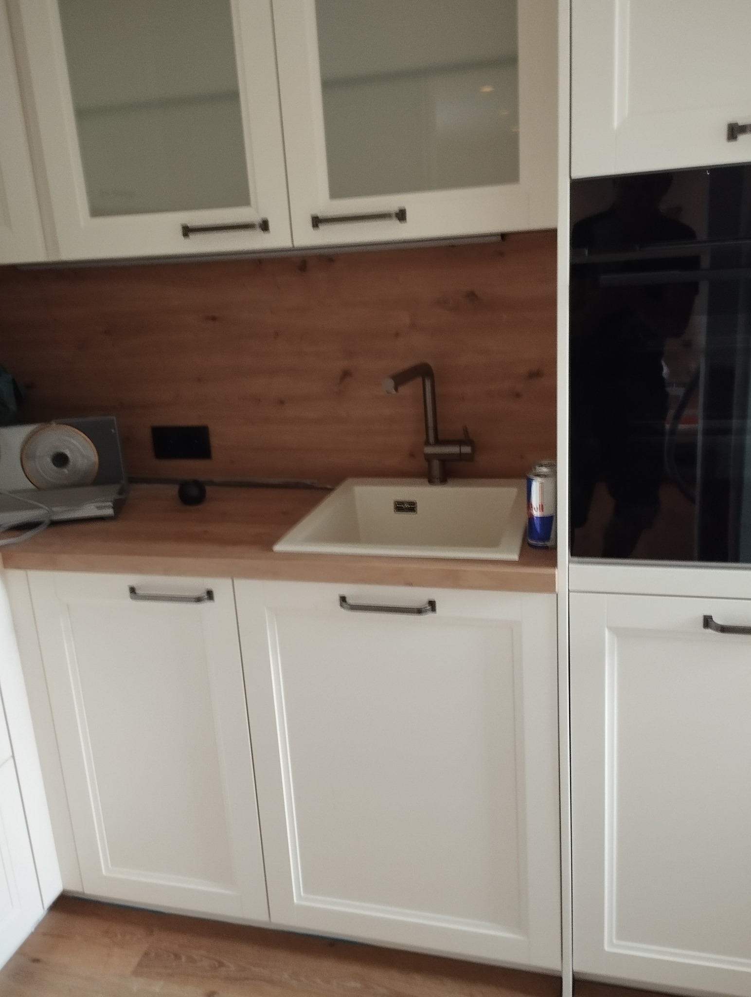 Nowoczesna kuchnia w bieli z drewnianym blatem i baterią przy zlewie. Widoczny fragment płyty indukcyjnej i piekarnika. Minimalistyczny design, ciepłe oświetlenie.