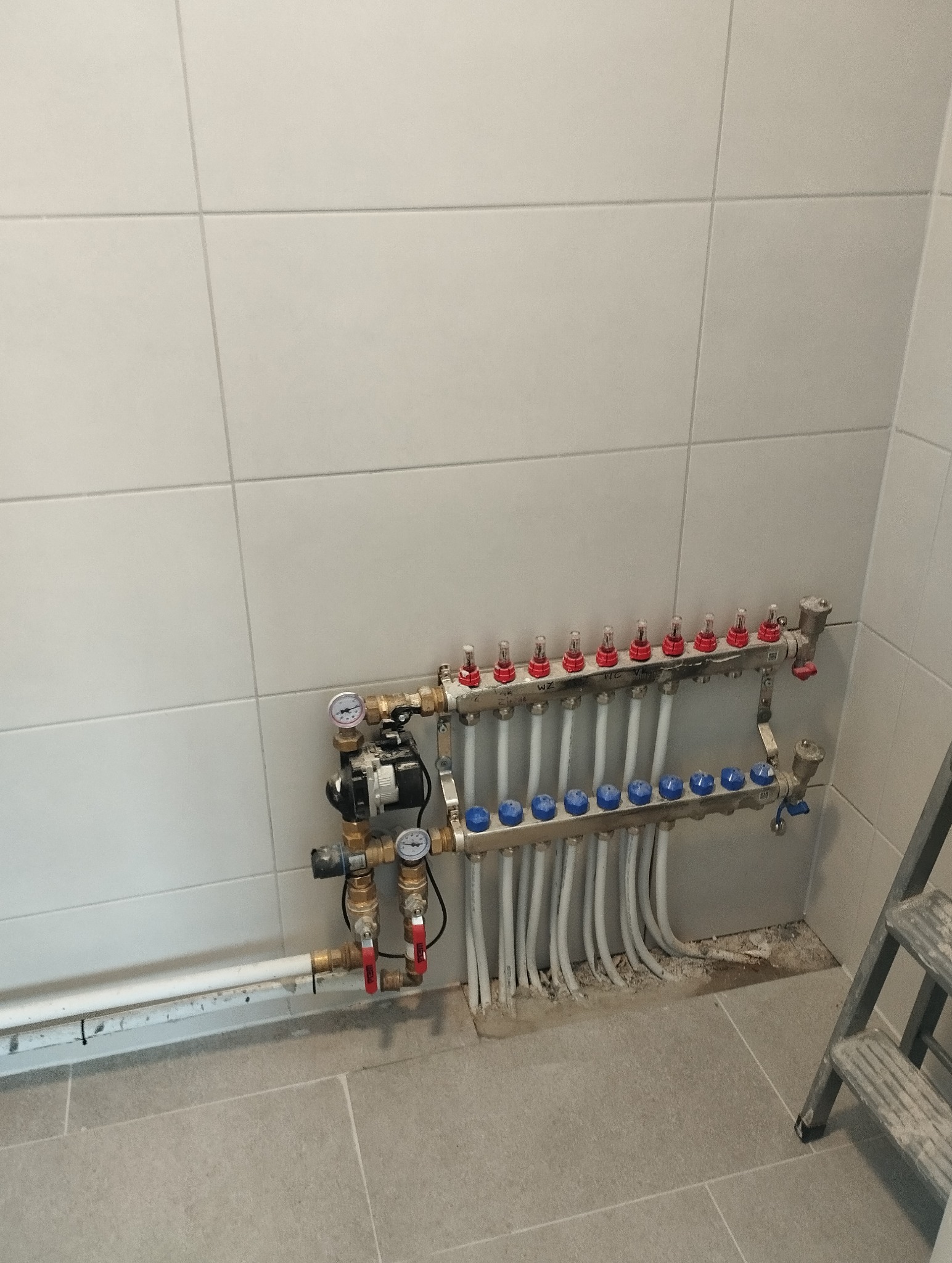 Rozdzielacz ogrzewania podłogowego z rurami na tle płytek. Widoczne manometry i zawory. Instalacja w łazience, drabina w prawym dolnym rogu.