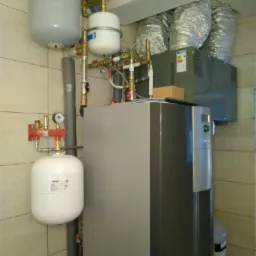 Szara pompa ciepła z widocznymi zbiornikami wyrównawczymi, rurami miedzianymi i elementami instalacji wentylacyjnej w pomieszczeniu z beżowymi płytkami.