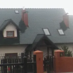 Dom jednorodzinny z szarym dachem pokrytym dachówką bitumiczną, widoczne okna dachowe, komin z metalowym wkładem oraz brama wjazdowa z ceglanymi słupkami.