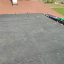 Czarna membrana EPDM ułożona na płaskim dachu z widocznymi narzędziami i elementami mocującymi, częściowo pokryta czerwoną dachówką, w tle pole i budynki.