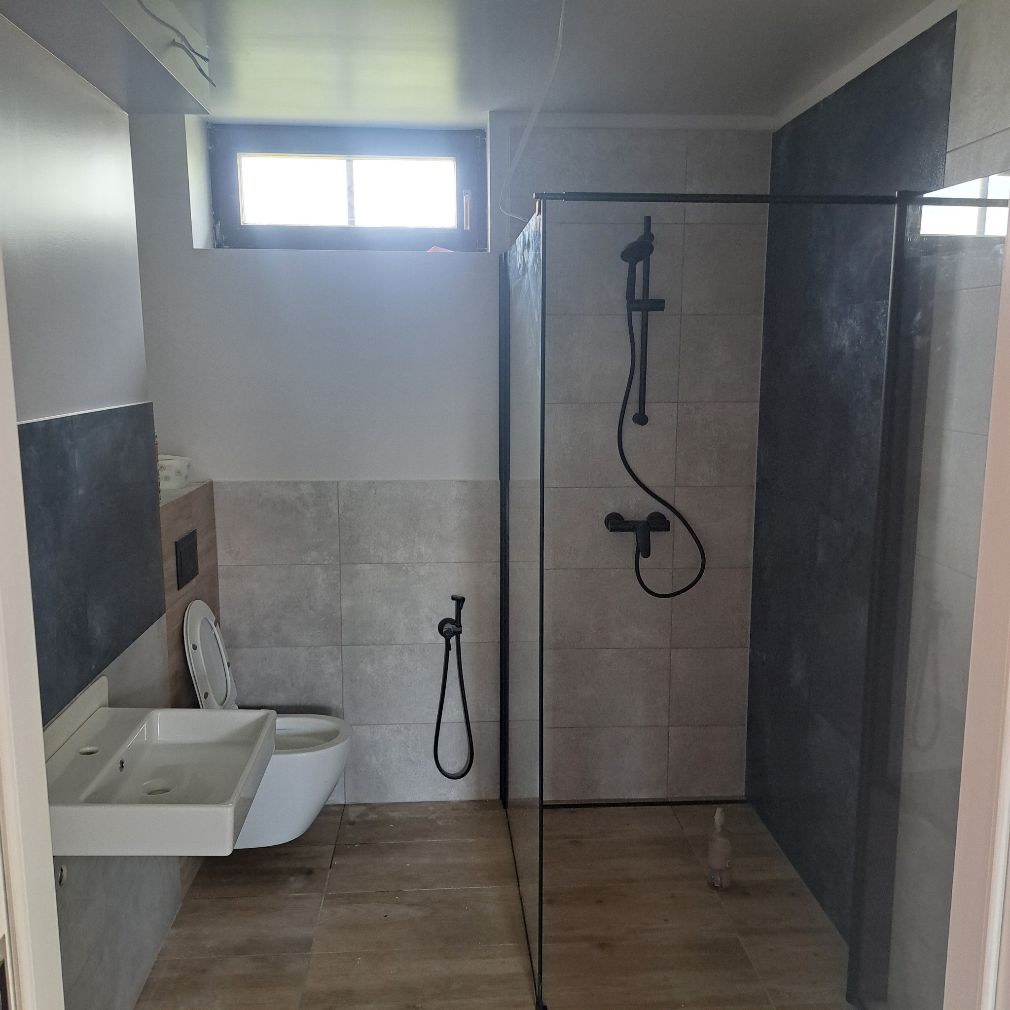 Nowoczesna łazienka z kabiną prysznicową typu walk-in, umywalką naścienną i toaletą. Dominuje szarość płytek i czarne akcenty armatury. Widoczne okno i drewnopodobna podłoga.