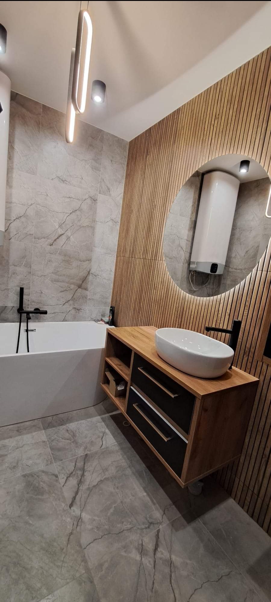Nowoczesna łazienka z drewnianą ścianą, owalną umywalką na szafce z szufladami i wanną. Lustro z podświetleniem LED odbija bojler. Szare płytki podłogowe.