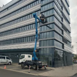 Wysięgnik koszowy na podwoziu ciężarówki, z pracownikiem w koszu na wysokości, przy szklanej fasadzie biurowca. Na pierwszym planie zaparkowany bus z reklamą montażu fasad aluminiowych.