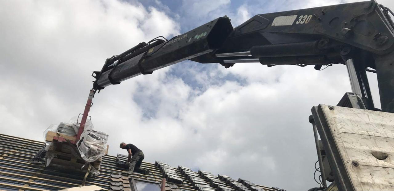 Żuraw samochodowy podnosi paletę dachówek na dach, gdzie pracuje dekarz przy montażu pokrycia dachowego.