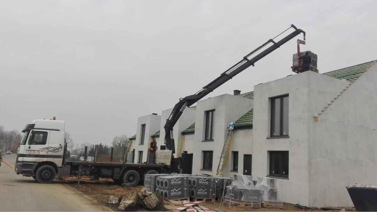 Ciężarówka z dźwigiem podnosi paletę z dachówkami na dach nowoczesnego domu w trakcie budowy, widoczne zapakowane dachówki na paletach na ziemi.