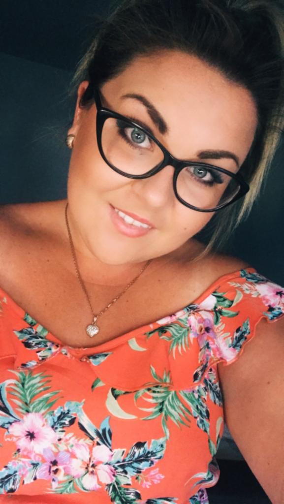 Portret uśmiechniętej kobiety w okularach i kwiecistej bluzce, z delikatnym naszyjnikiem w kształcie serca, na ciemnym tle. Ujęcie z bliska.