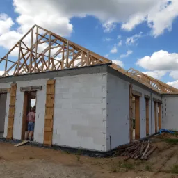 Budowa domu z białych bloczk&oacute;w: ściany wzniesione, drewniana konstrukcja dachu w trakcie montażu, widoczna osoba wewnątrz budynku. Niebo z chmurami w tle.