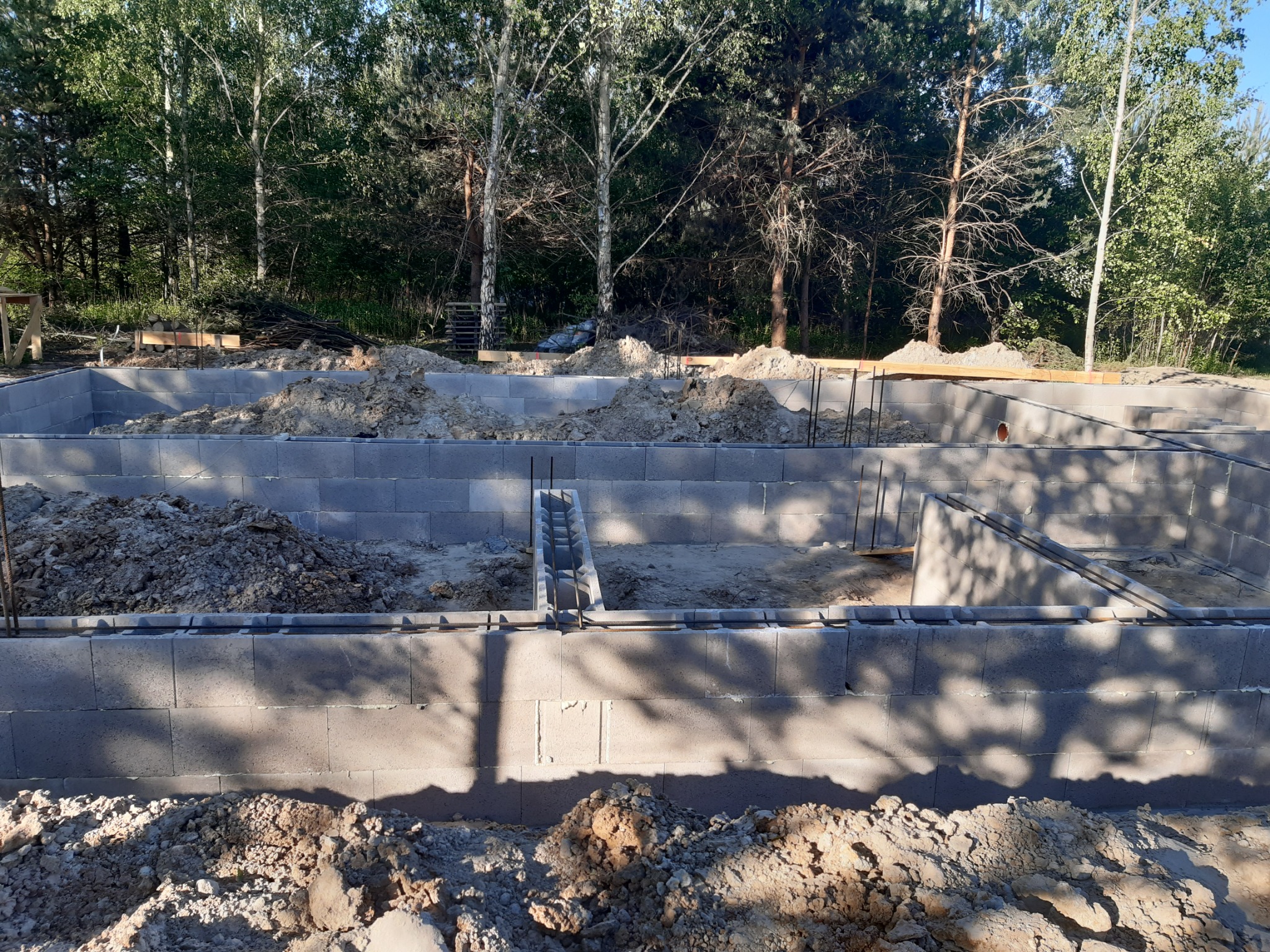 Wykopane fundamenty pod budynek, z widocznymi bloczkami betonowymi tworzącymi ściany, częściowo zasypane ziemią, z lasem w tle.