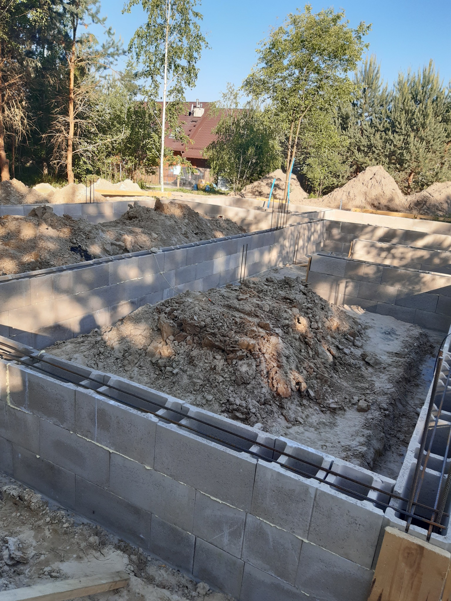 Fundamenty domu z bloczków betonowych, zbrojone stalą, z widoczną stertą ziemi wewnątrz wykopu i fragmentem domu w tle