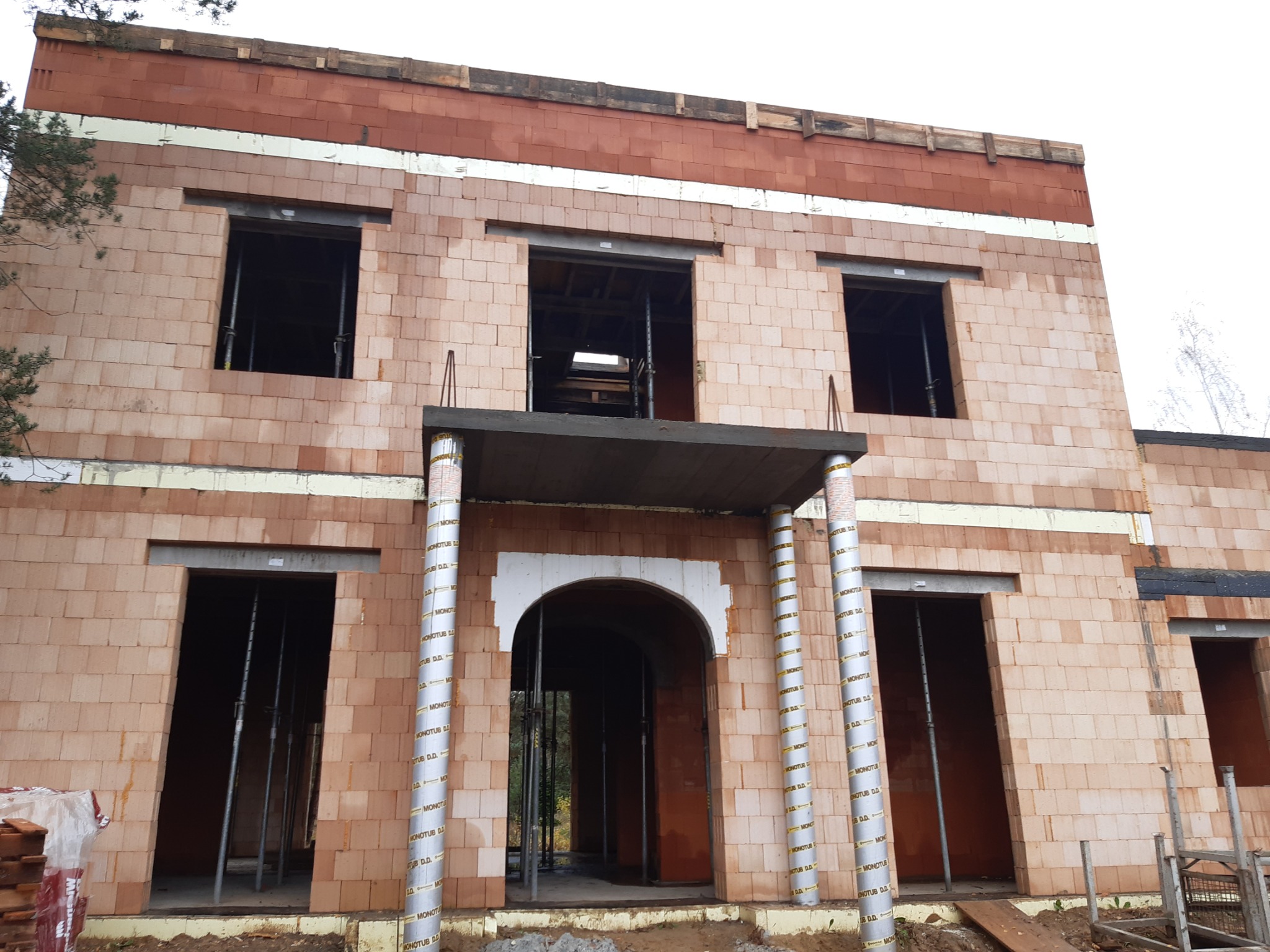 Wznoszony budynek z czerwonej cegły ceramicznej Porotherm, widoczne otwory okienne i drzwiowe, podparte stemple budowlane MONOTUB D.D., otynkowane nadproża, brak dachu.