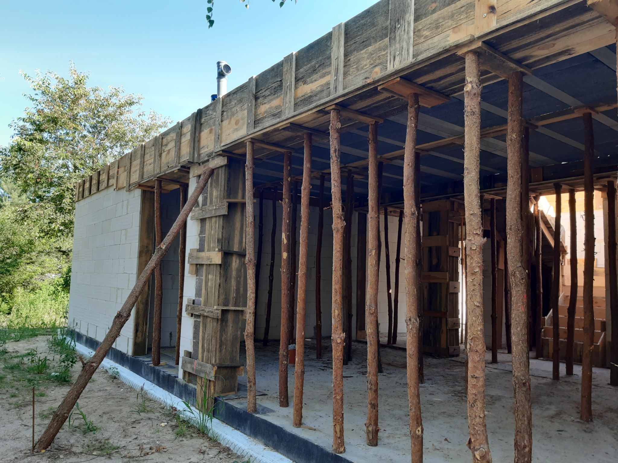 Surowy stan budowy domu murowanego z betonu komórkowego, widoczne drewniane szalunki i stemple podtrzymujące strop, fragment dachu z kominem, otoczenie w stanie naturalnym.