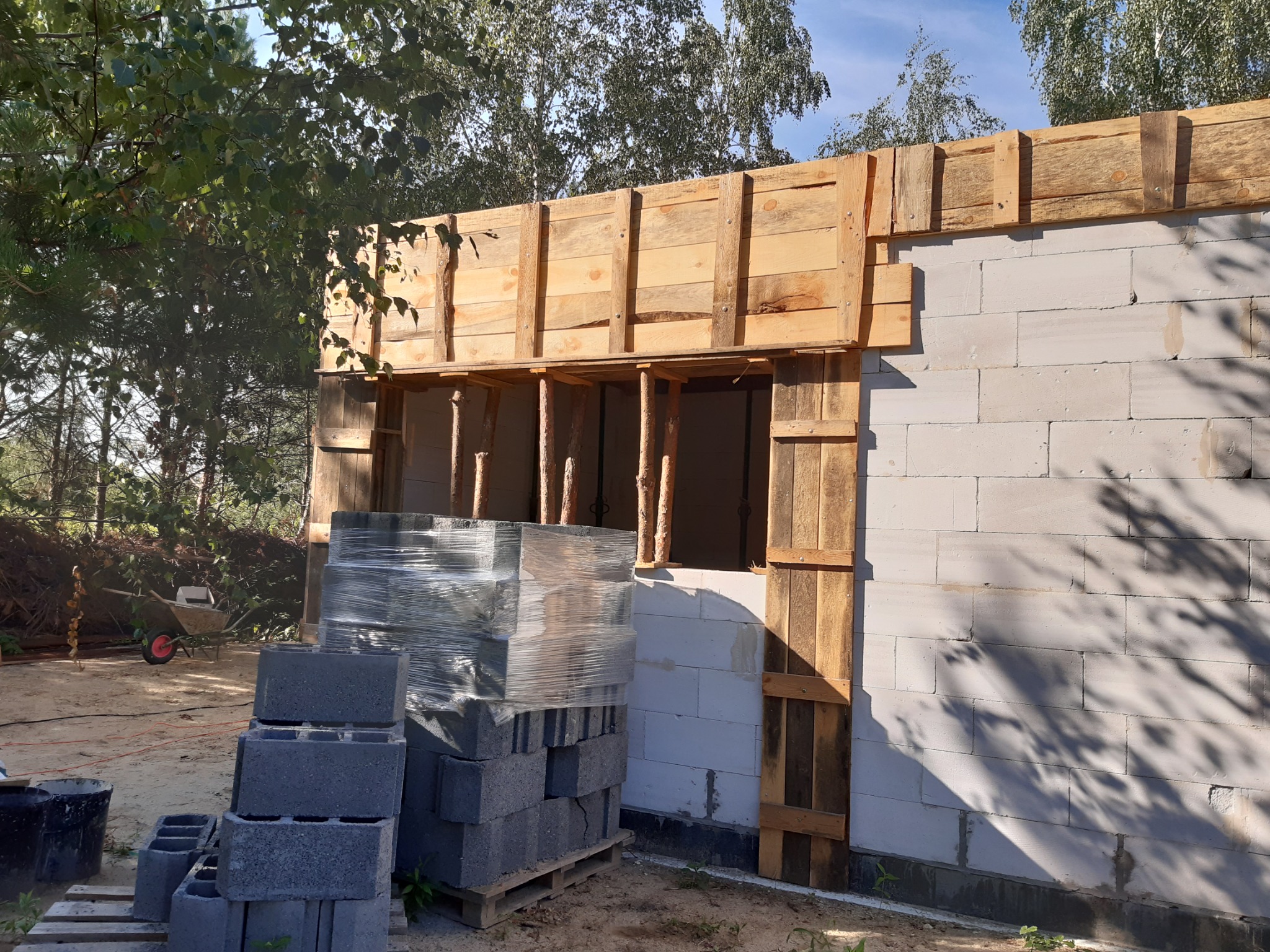 Wznoszona ściana budynku z betonu komórkowego z drewnianym szalunkiem nad otworem okiennym, paleta z bloczkami budowlanymi w folii obok ściany, taczka w tle.
