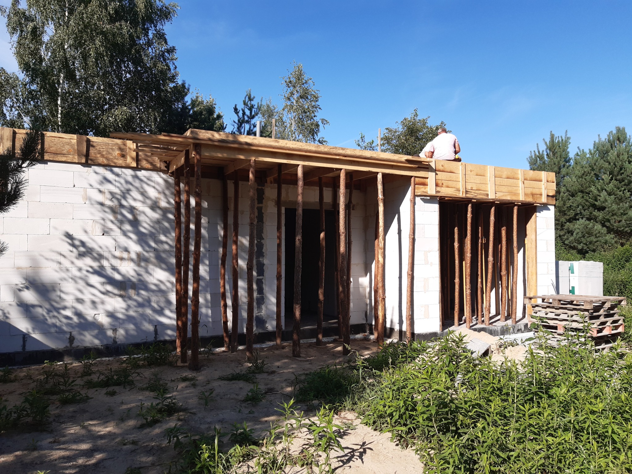 Wznoszenie ścian parterowego budynku z białych bloczków, z drewnianym szalunkiem i podporami, widoczny pracownik na szalunku, teren budowy z roślinnością.