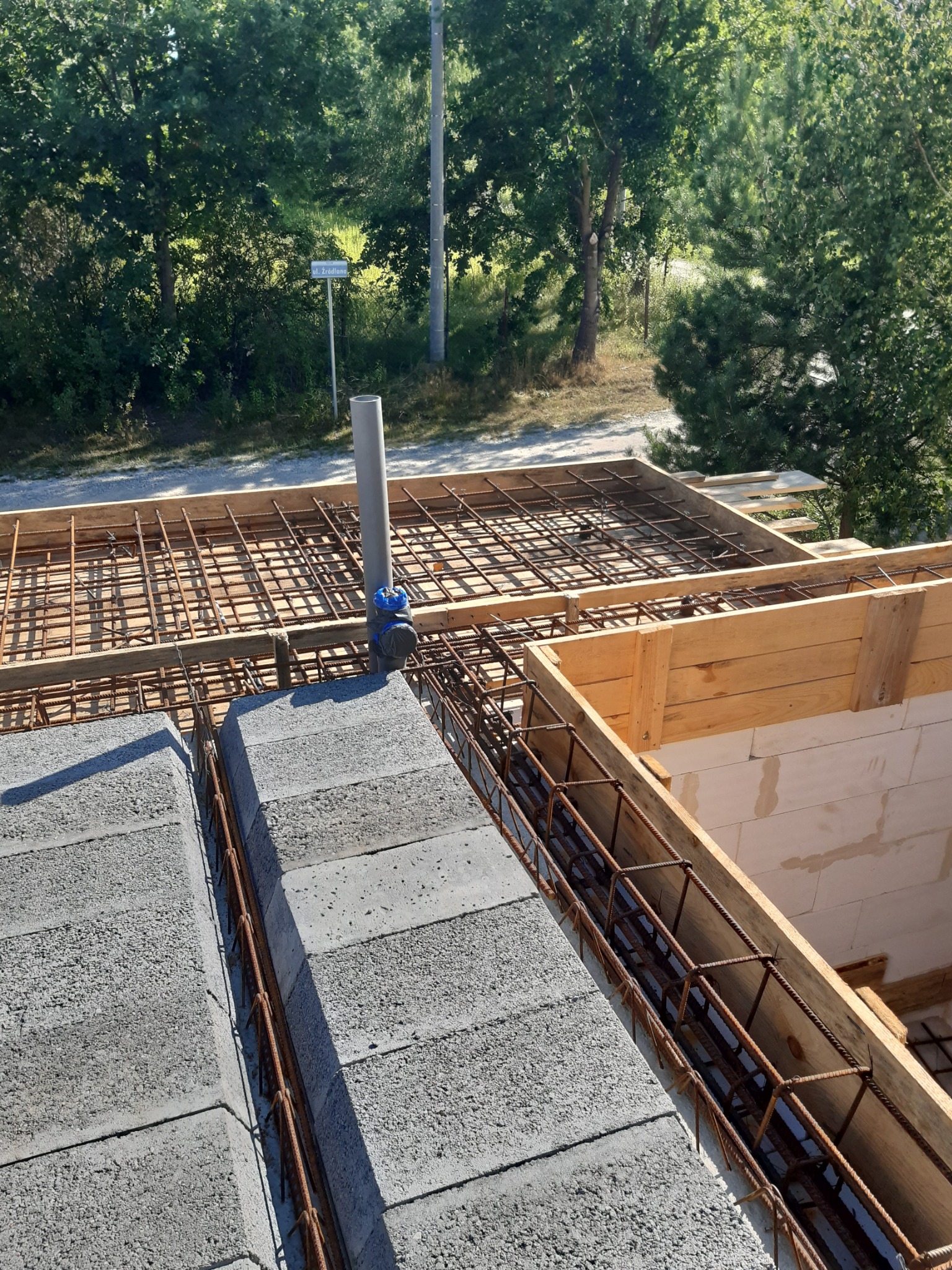 Wznoszenie ścian fundamentowych z bloczków betonowych, widoczne zbrojenie i szalunek, w tle zieleń drzew i znak drogowy.