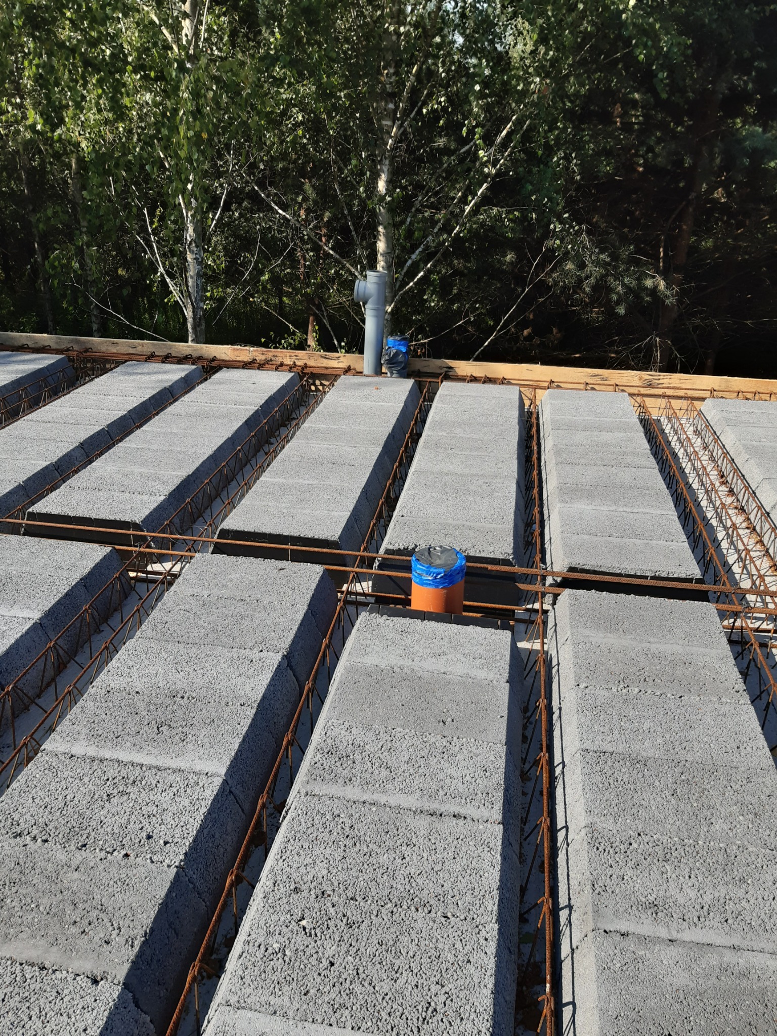 Szare bloczki stropowe ułożone na konstrukcji, z widocznym zbrojeniem i pionowymi rurami instalacyjnymi, na tle drzew.