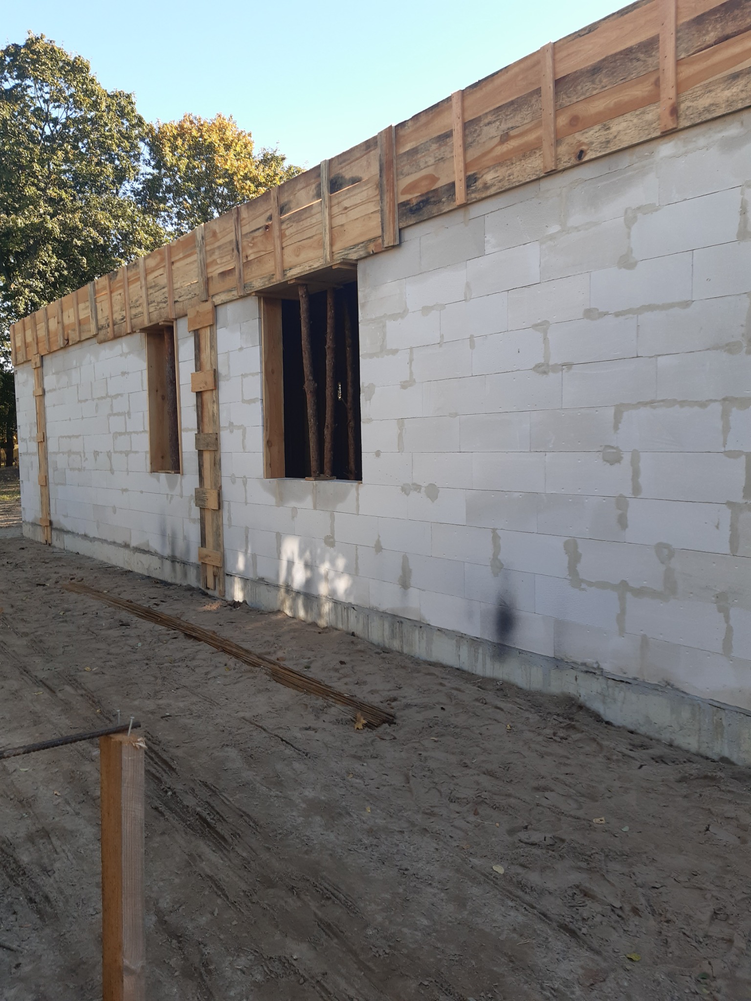 Wznoszenie ścian parterowego budynku z bloczków betonowych, widoczne drewniane szalunki okienne i obwodowe, teren budowy z ziemią i wystającymi prętami zbrojeniowymi.