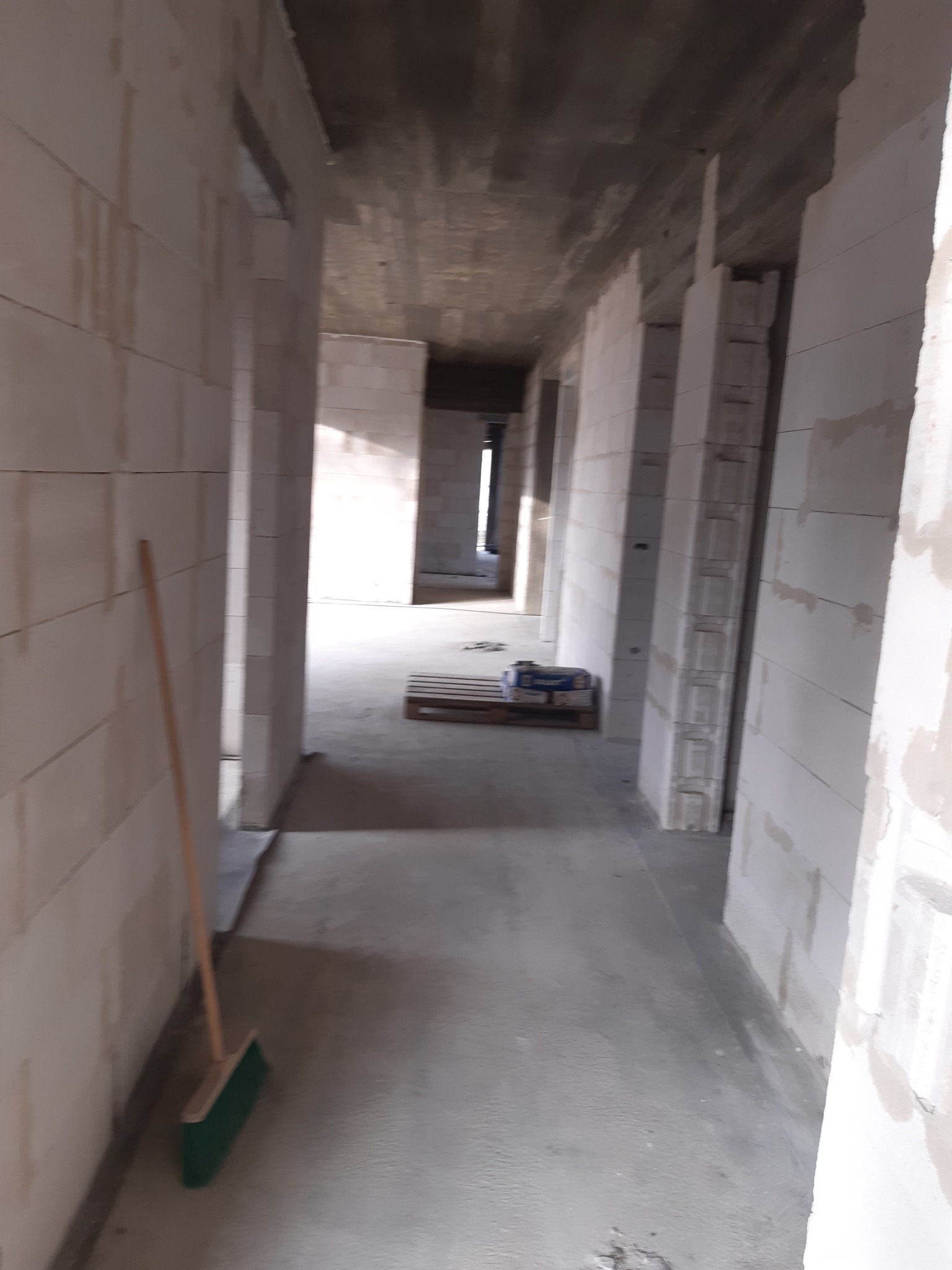 Wnętrze w trakcie budowy z białymi ścianami z betonu komórkowego, surowym betonowym sufitem, widoczną paletą z workami cementu i zieloną zmiotką opartą o ścianę.