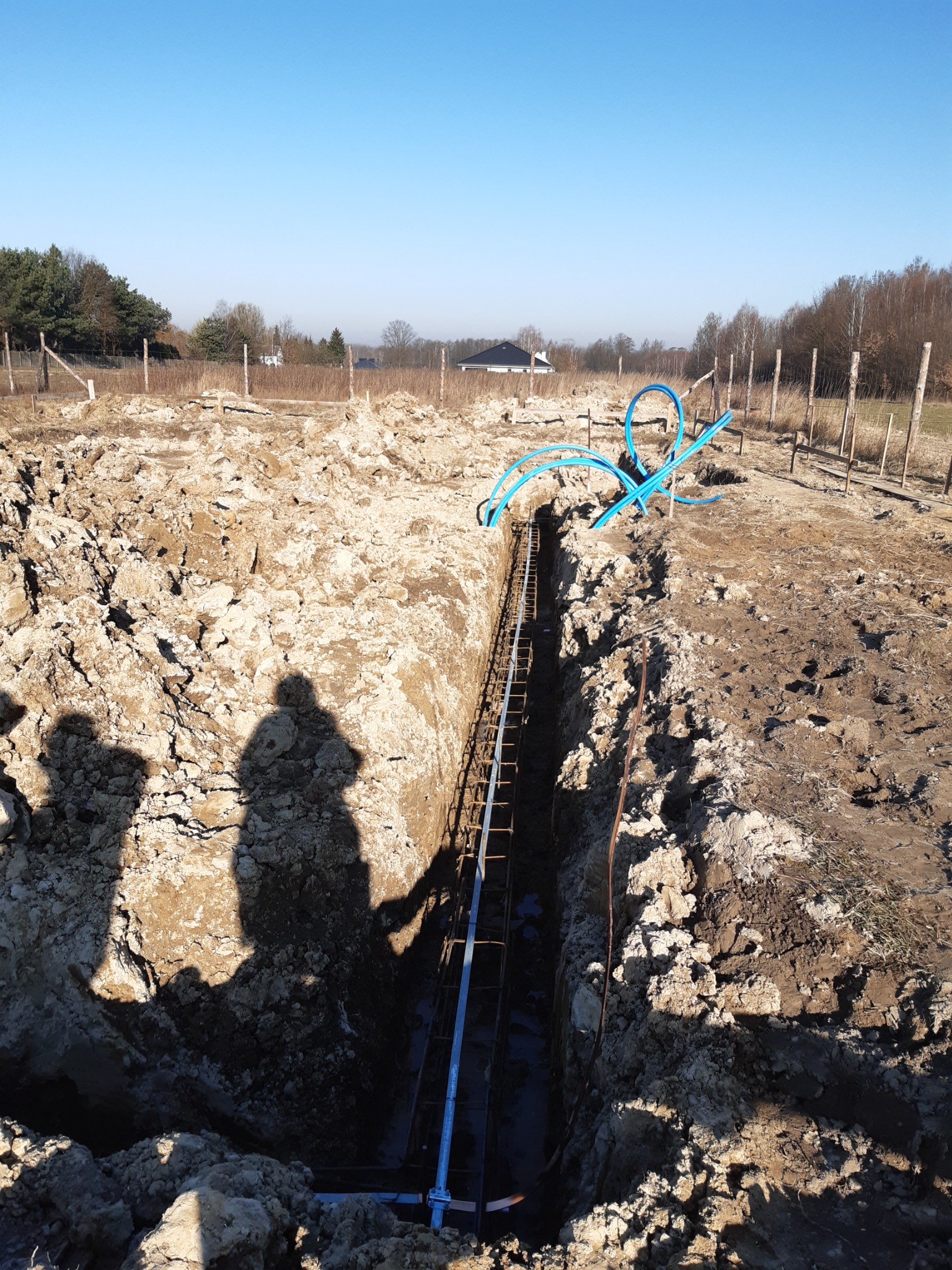 Wykop pod fundamenty z ułożonym zbrojeniem i niebieskimi rurami instalacyjnymi, widoczne cienie dwóch osób oraz odległy krajobraz z domem.
